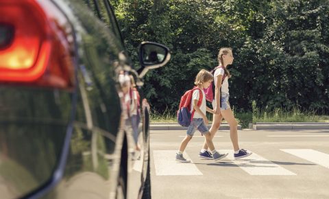 Ein Mädchen geht mit ihrem jüngeren Bruder über einen Zebrastreifen. Sie hält den Jungen an der Hand, Im Vordergrund sieht man im Anschnitt ein schwarzes Auto.