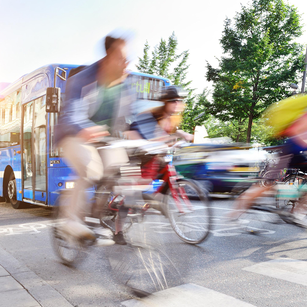 Fahrradfahrende auf einer belebten Straße, ein Bus im Hintergrund. Leichte Unschärfe gibt dem Bild Bewegung.