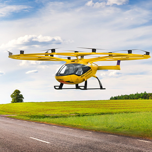 Bild eines Multikopter in der Luft. Er fliegt über einer Straße, im Hintergrund grüne Wiese und bewölkter Himmel.