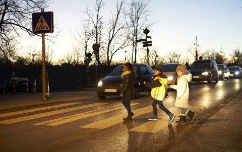 Drei Kinder überqueren in der Dämmerung einen Zebrastreifen: Eines trägt helle Kleidung, eines dunkle Kleidung und eines eine Warnweste.