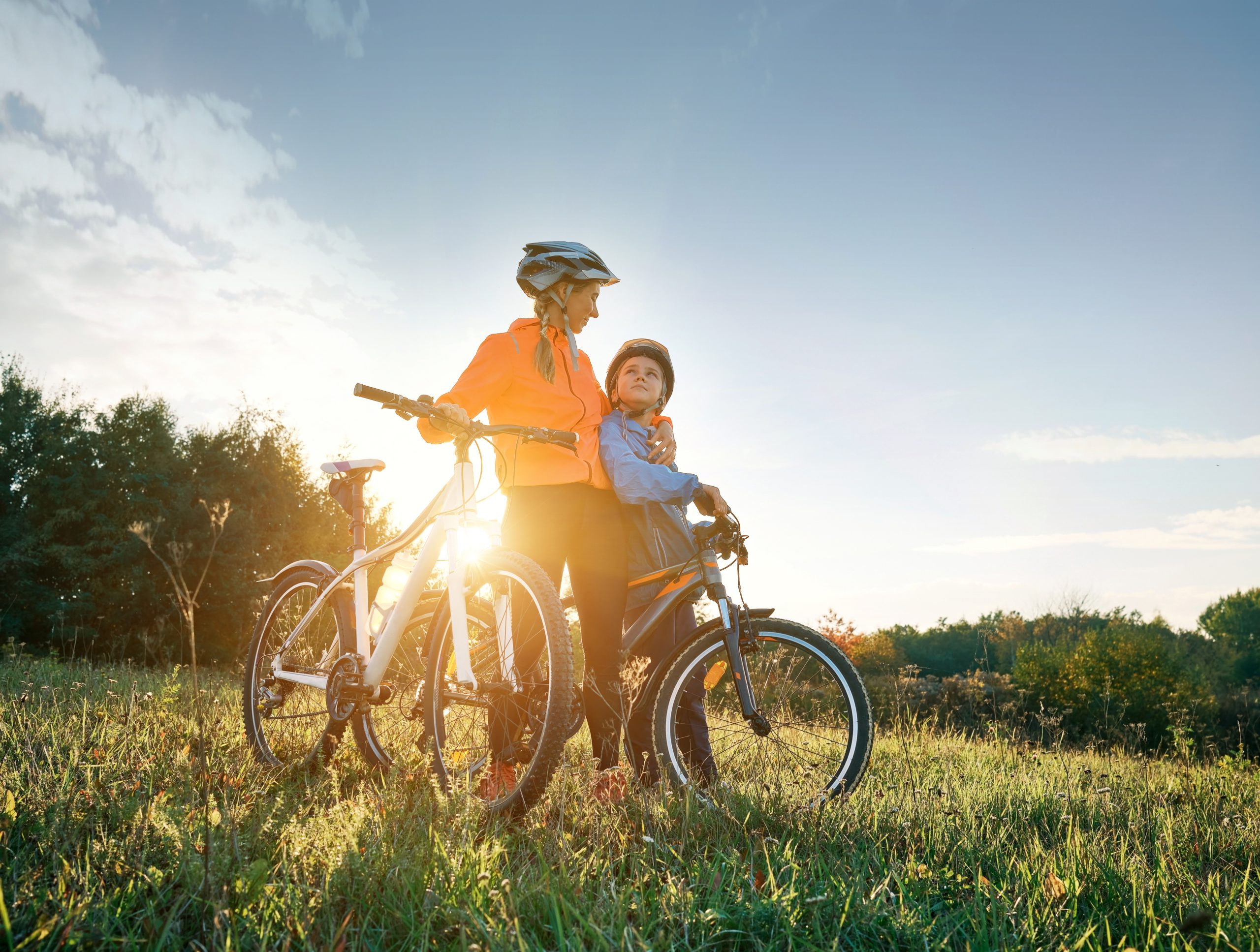 Mutter und Sohn fahren Fahrrad. Glücklicher Junge mit Helm neben seiner Mutter auf einer grünen Wiese an einem Herbsttag bei Sonnenuntergang. Mutter und Kind sind zusammen aktiv in der Natur.