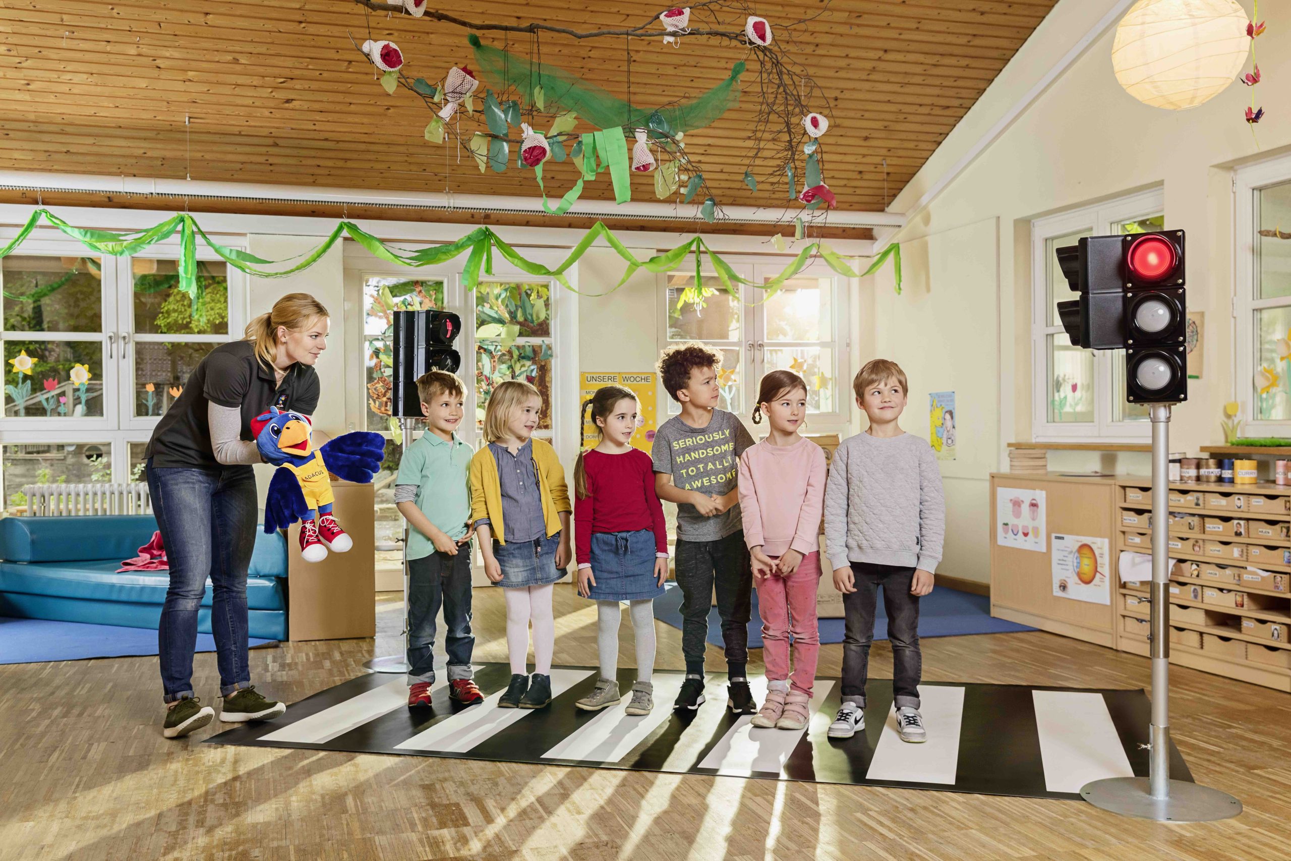 Eine junge Frau steht in einem Gruppenraum einer Kita und hält eine Handpuupe, den Raben ADACUS, in der Hand. Sie erklärt den Kindern als Trainerin, wie man sicher über eine Ampel und einen Zebrastreifen geht. Kinder stehen auf einem Zebrastreifen, rechts im Bild steht eine Übungsampel.