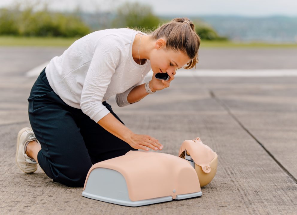 Eine junge Frau kniet vor einer Reanimationspuppe. Sie hält ein Handy in ihrer Hand.