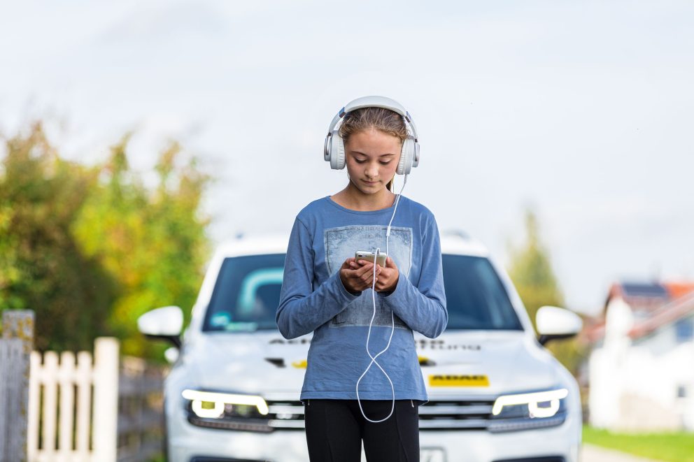 Ein Mädchen steht auf der Straße, sie hat Kopfhörer auf und schau auf ihr Handy. Hinter ihr nähert sich ein Auto.