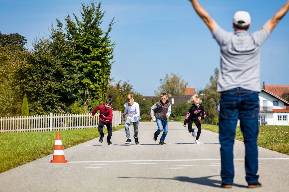 4 Kinder sprinten auf einer Straße los. Ein Moderator steht im Vordergrund des Bildes, neben ihm ein Verkehrshütchen.