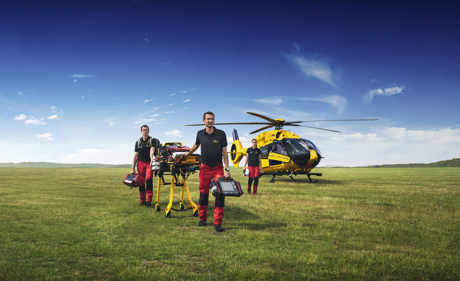 Mitarbeitende der ADAC Luftrettung auf einer Wiese, sie schieben eine Trage. Der gelbe Helikopter steht im Hintergrund. Blauer Himmel.