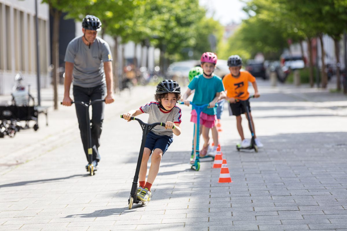 3 Kinder und ein Mann mit Helm auf Rollern. Sie fahren um Hütchen in einer Fußgängerzone.