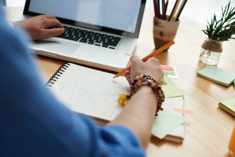 Im Anschnitt ist ein Laptop zu sehen und eine Person, die auf einem Block Notizen macht.