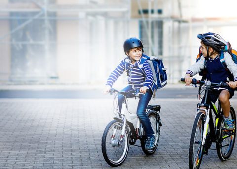 Ein Junge mit Helm und Schulranzen auf einem Fahrrad, er unterhält sich mit einem anderen Kind, welches ebenfalls Fahrrad fährt, im Anschnitt zu sehen.