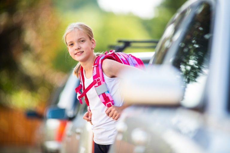 Ein Mädchen auf dem Heimweg nach der Schule. Sie geht zu Fuß und schaut zwischen parkenden Autos hervor.