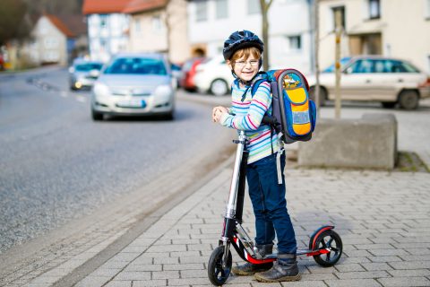 Ein Junge steht mit seinem Roller an einer Straße. Er trägt einen Schulranzen und Helm. Im Hintergrund ein heranfahrendes Auto.