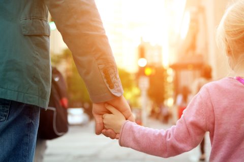 Ein Vater geht mit seiner Tochter Hand in Hand. Im Ausschnitt des Bildes sieht mal nur die Arme und Hände. Im Hintergrund eine Straße, freundliches sonniges Licht