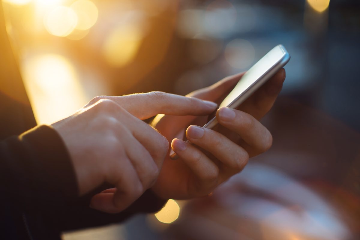 Ein Mann hält ein Smartphone in seinen Händen. Das Bild zeigt eine Nahaufnahme des Smartphones und der Hände. Im Hintergrund ein softes Sonnenlicht.