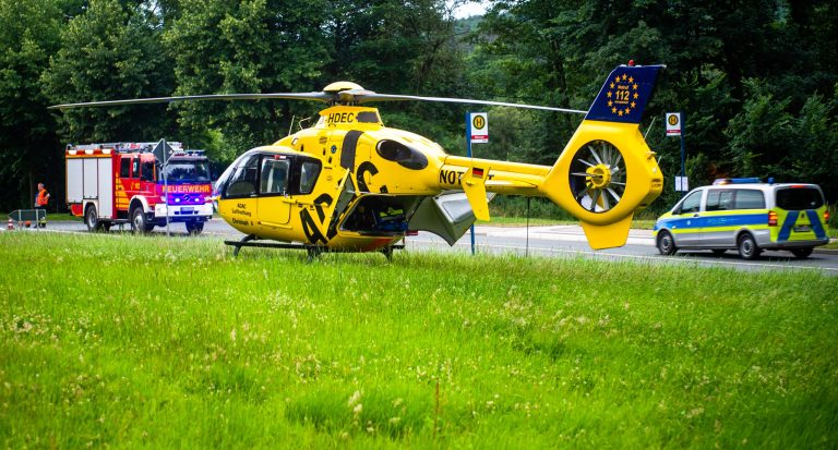 Ein gelber Rettungshubschrauber der ADAC Luftrettung steht auf einer Grasfläche nahe einer Straße. Im Hintergrund sind Einsatzfahrzeuge, darunter ein rotes Feuerwehrauto und ein Polizeiwagen mit blauen und gelben Markierungen, zu sehen. Mehrere Einsatzkräfte sind in der Nähe der Fahrzeuge sichtbar, und Bäume säumen den Hintergrund.