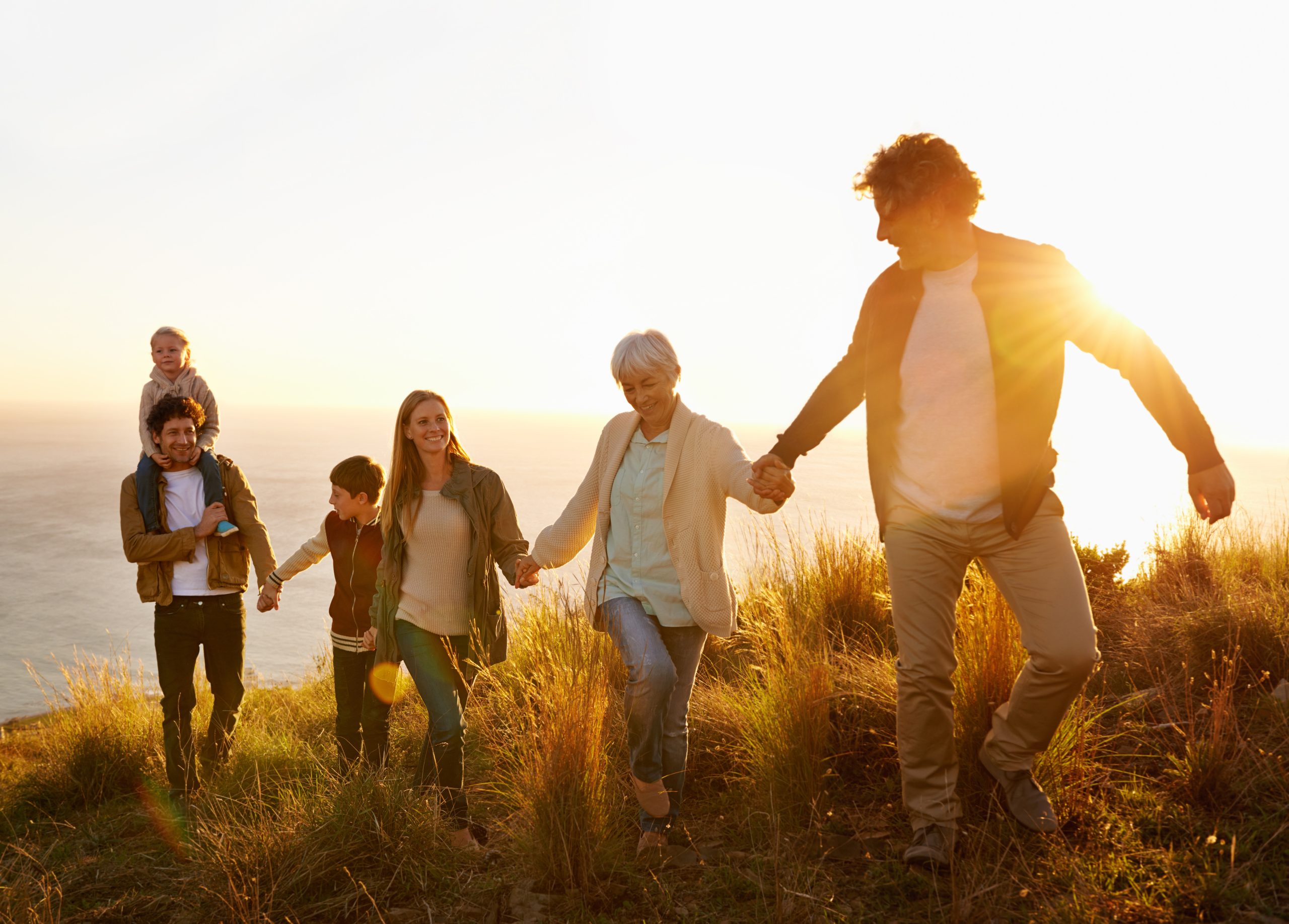 Eine Familie wandert glücklich auf einem grünen Hügel an der Küste, im Hintergrund das Meer – Mehrgenerationenmoment mit Kind, Eltern und Großeltern im Sonnenlicht. Ein idyllisches Bild einer Familie, die zusammen die Natur genießt, während die Sonne sanft auf die Szene scheint.