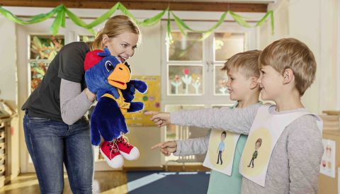 Eine Frau hält eine blaue Handpuppe in der Hand, den Raben ADACUS. Die Frau ist eine Moderatorin und spricht den Raben. 2 Jungen lächeln und zeigen auf den Raben. Sie befinden sich in einem Gruppenraum eines Kindergartens.