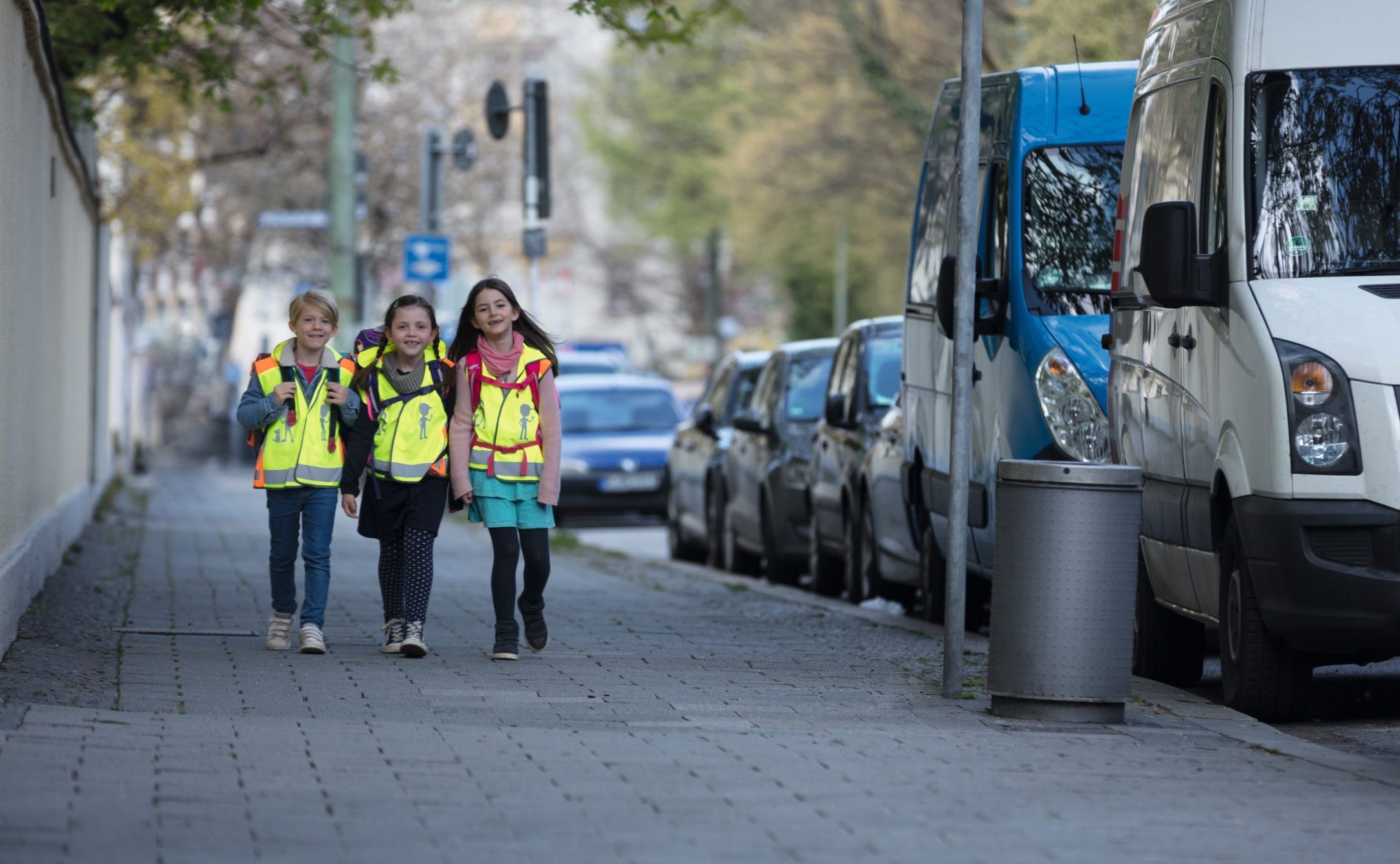 Zwei Mädchen und ein Junge im Grundschulalter gehen auf einem Gehweg in der Stadt. Sie tragen gelbe Sicherheitswarnwesten und Schulranzen. Ihr Stimmung ist gut, sie lachen und haben Freude gemeinsam auf dem Schulweg.