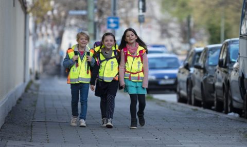 Zwei Mädchen und ein Junge im Grundschulalter gehen auf einem Gehweg in der Stadt. Sie tragen gelbe Sicherheitswarnwesten und Schulranzen. Ihr Stimmung ist gut, sie lachen und haben Freude gemeinsam auf dem Schulweg.