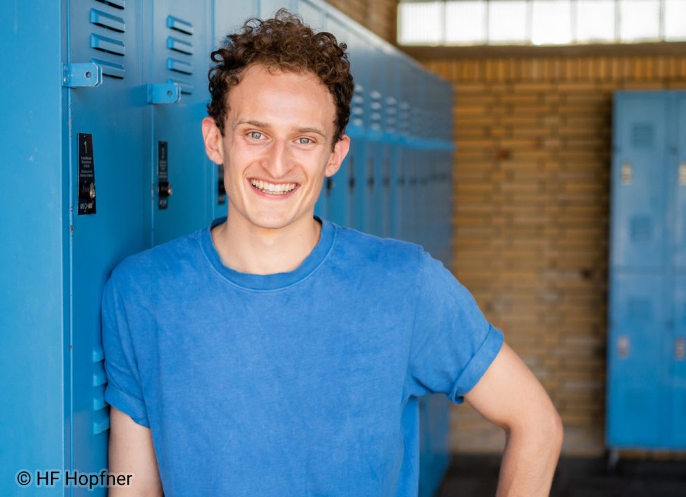 Julian Janssen, bekannt als Checker Julian, in einem blauen T-Shirt, lächelnd an einem blauen Spint lehnend.