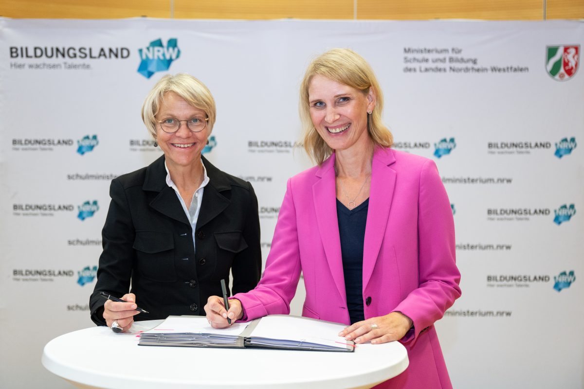 Vorständin der ADAC Stiftung Christina Tillmann und Dorothee Feller, Ministerin für Schule und Bildung des Landes Nordrhein-Westfalen, unterzeichnen ein Dokument. Sie stehen gemeinsam in die Kamera lächelnd an einem Pult.