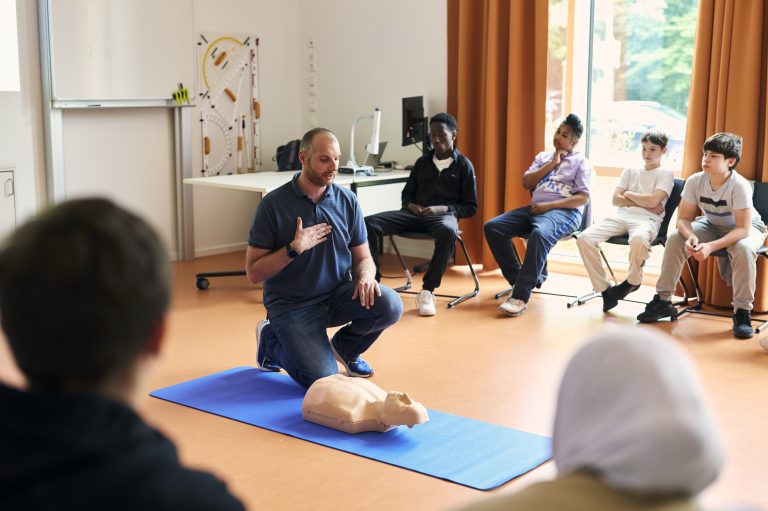 Ein Lehrer kniet in der Mitte eines Klassenzimmers. Vor ihm liegt eine Reanimationspuppe. Er schult Kinder, die im Kreis um ihn herum auf Stühlen sitzen, im Reanimationsunterricht.