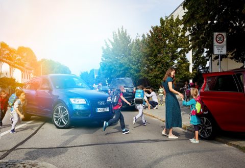 Verkehrschaos vor einer Grundschule. Kinder laufen über die Straße, während Autos auf der Straße wenden und auf Gehwegen parken.