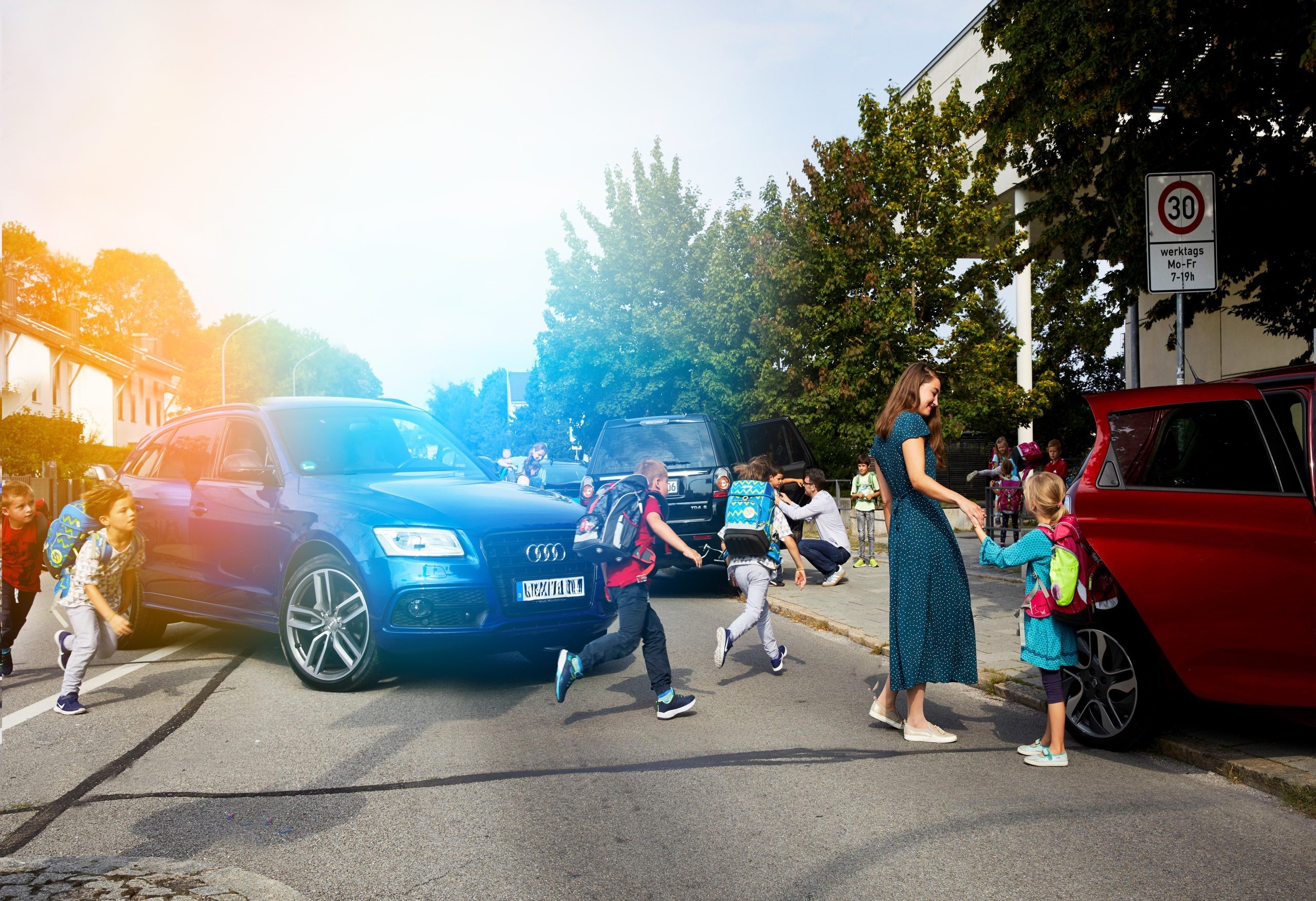 Verkehrschaos vor einer Grundschule. Kinder laufen über die Straße, während Autos auf der Straße wenden und auf Gehwegen parken.