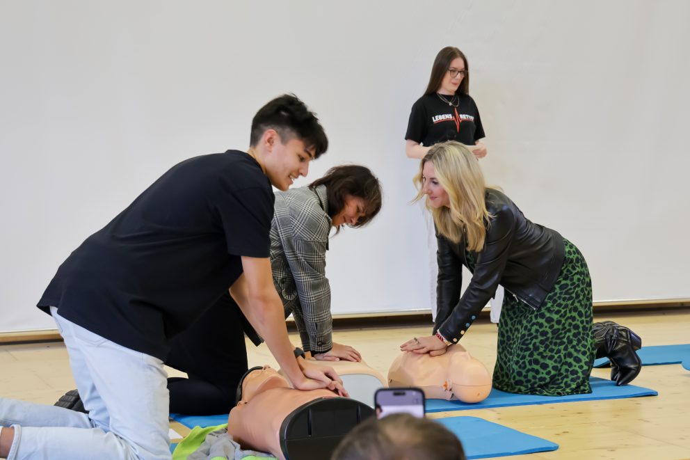 Kultusministerin Anna Stolz übt in einer Turnhalle an einer Reanimationspuppe die Wiederbelebung. Neben ihr führen weitere Teilnehmende der Fortbildungsoffensive zur Einführung von Reanimationsunterricht in Bayern die Übung durch.