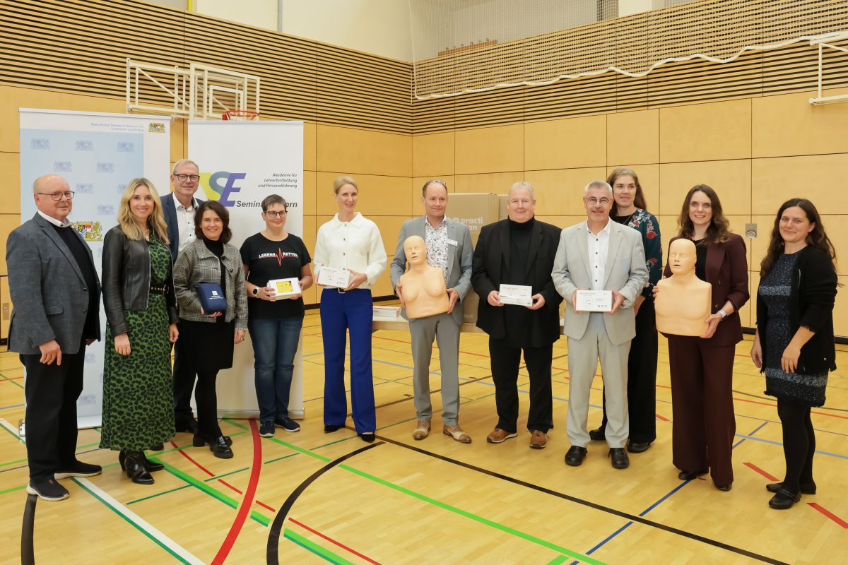 Gruppenfoto in einer Turnhalle mit Teilnehmenden der Fortbildungsoffensive zur Einführung von Reanimationsunterricht in Bayern. Unter den Anwesenden sind Kultusministerin Anna Stolz und Christina Tillmann, Vorständin der ADAC Stiftung. Einige Personen halten Reanimationspuppen in den Händen.