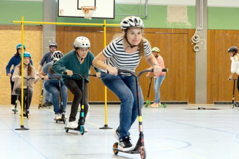 Lehrkräfte üben in der Turnhalle den sicheren Umgang mit dem Tretroller im Rahmen des Programms „Roller Fit“.