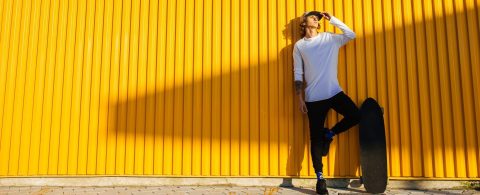 Ein hipper, junger Mann steht mit seinem Longboard vor einer gelben Wand.