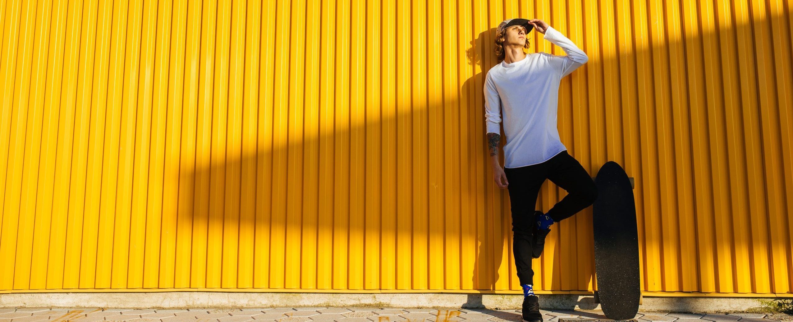 Ein hipper, junger Mann steht mit seinem Longboard vor einer gelben Wand.