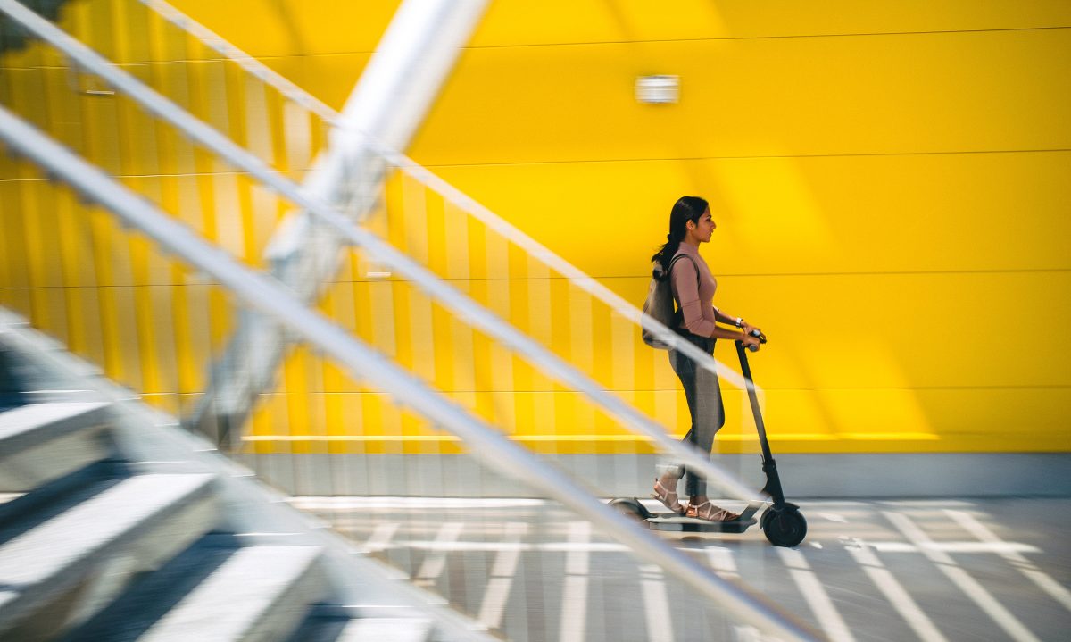 Eine junge Frau fährt auf einem E-Roller vor einer gelben Wand. Treppe und Boden im Vordergrund sind leicht unscharf, was dem Bild eine dynamische Wirkung verleiht.