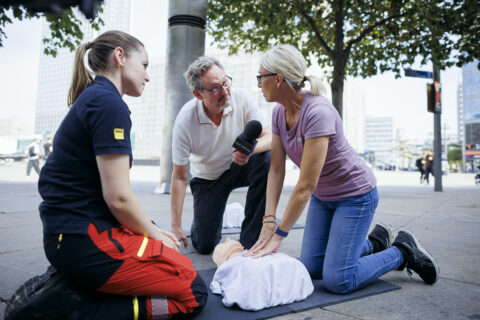 Dr. Eckart von Hirschhausen, eine Passantin und Notfallsanitäterin Katharina Helm im Gespräch auf dem Alexanderplatz in Berlin. Sie knien neben einer Reanimationspuppe auf dem belebten Platz, während im Hintergrund Passantinnen und Passanten sowie umliegende Gebäude zu sehen sind.
