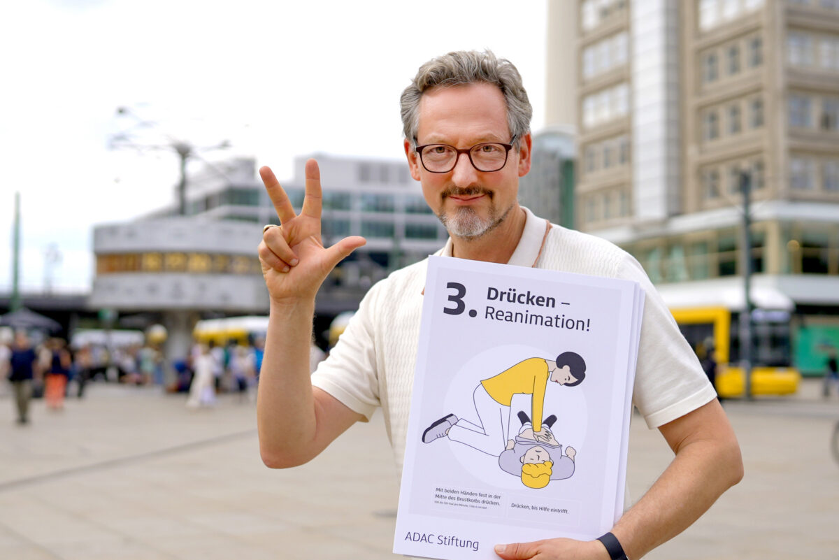 Eckart von Hirschhausen steht auf einem öffentlichen Platz und hält ein Schild mit dem dritten Wiederbelebungsschritt „Drücken“ hoch. Mit drei ausgestreckten Fingern verweist er auf den dritten Schritt der Wiederbelebung.