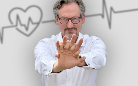 Porträt von Dr. Eckart von Hirschhausen mit nach vorne ausgestreckten Armen. Die übereinandergelegten Hände zeigen die Handhaltung für die Herzdruckmassage. Im Hintergrund sind ein stilisiertes Herz und eine Herzschlaglinie dargestellt.