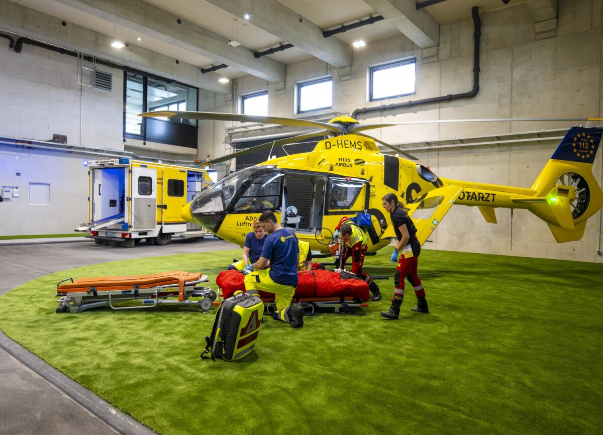 Helikopter der ADAC Luftrettung in einer Übungshalle, mit Crew im Vordergrund. Übungs-Rettungswagen im Hintergrund.